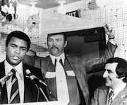 Heavyweight champion Muhammad Ali spoke at a news conference in Cleveland on Feb. 11, 1975, and reiterated his statement that his profits from his championship fight with Chuck Wepner would go helping black causes. Wepner stands behind the champion holding a banner and Nick Mileti, who owns the Coliseum where the fight will be held on March 24, looks on. (AP Photo/Barry Thumma)